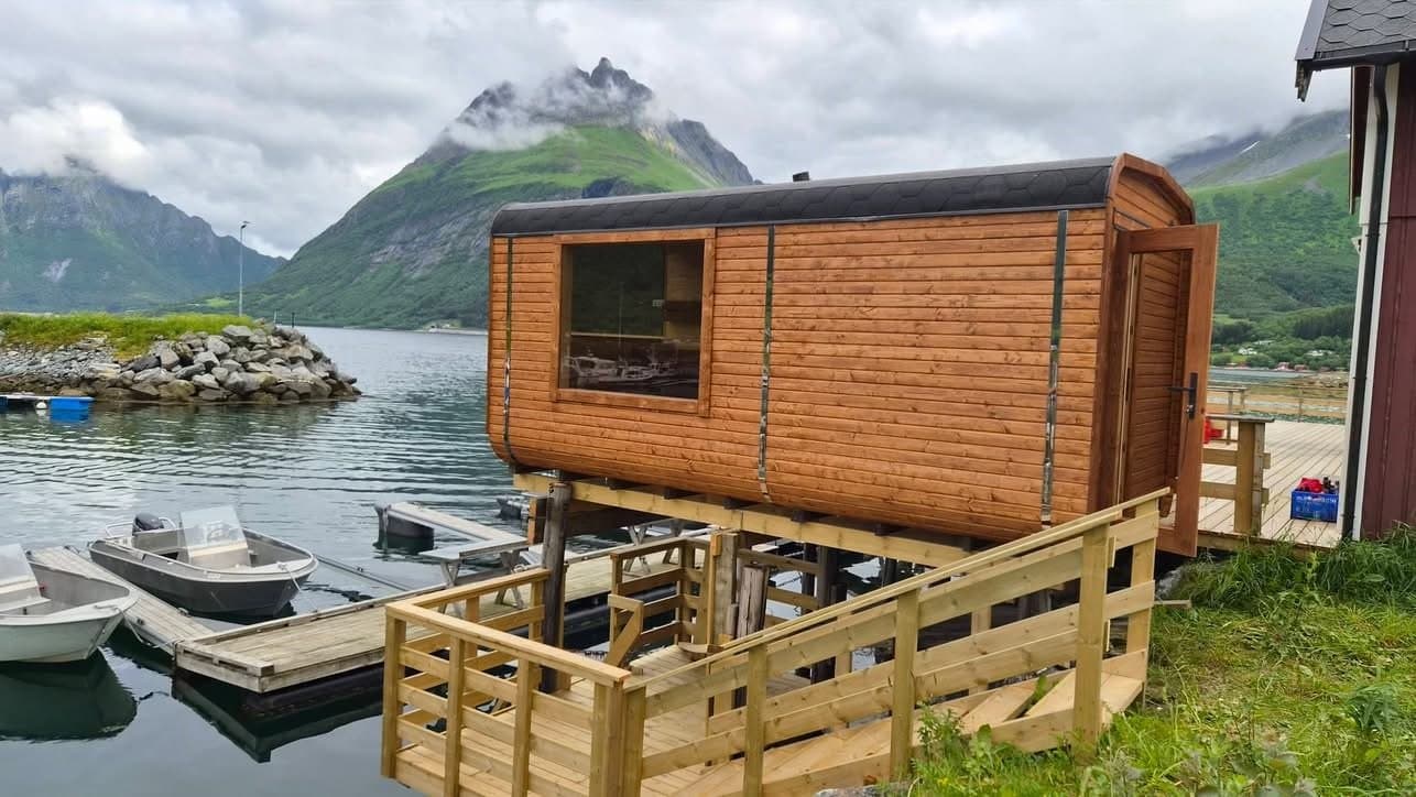 Badstue med terrasse og trapp på en brygge ved sjøen, med båter og fjell bak