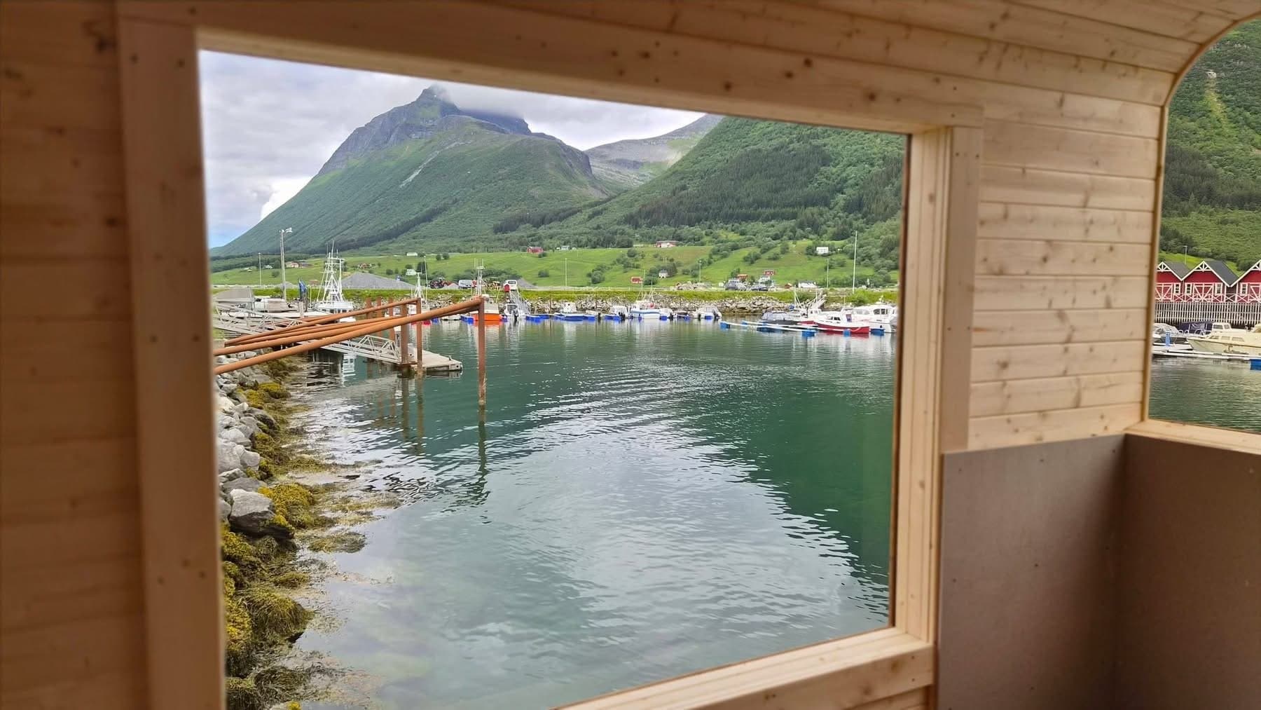 Utsikt gjennom vinduet i badstuen mot en småbåthavn, røde sjøhus og grønne fjell
