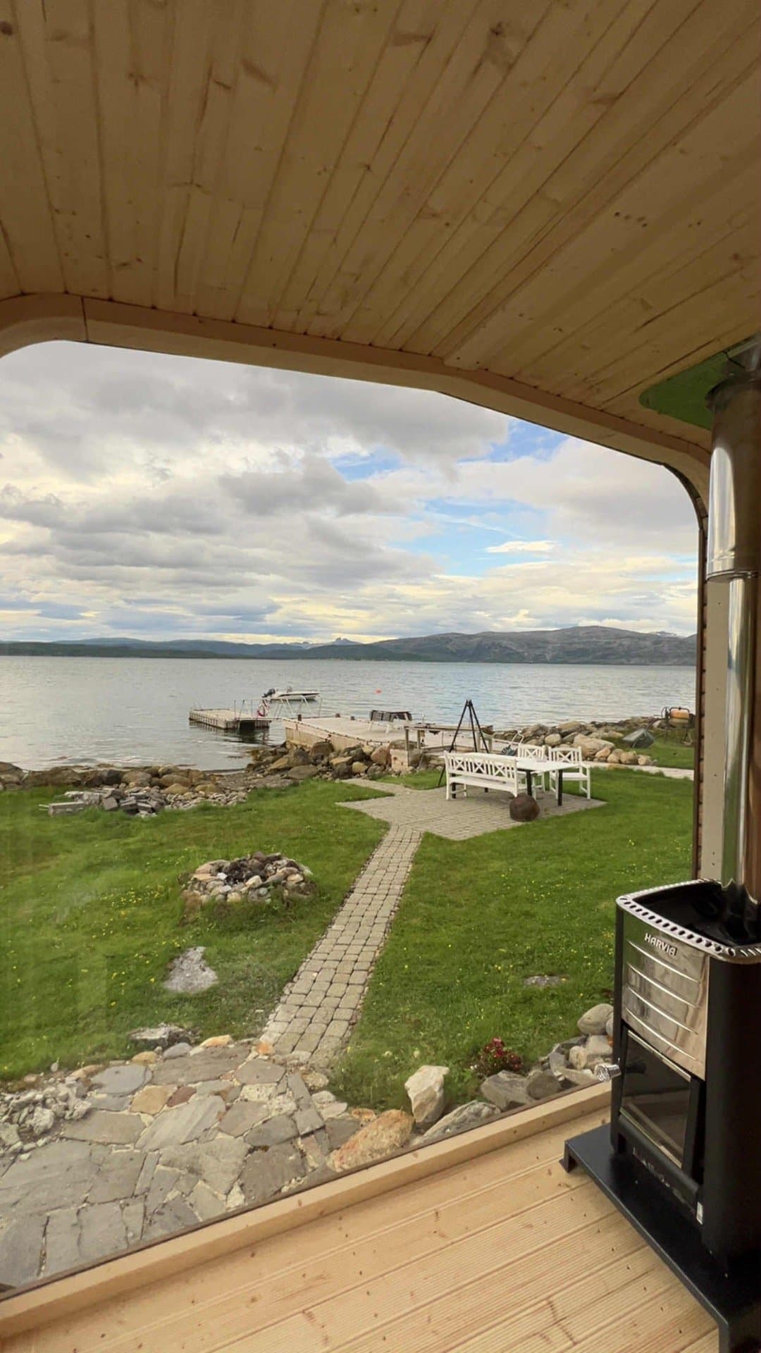Utsikt fra badstuen gjennom glassfronten mot en hage med steinsti, en brygge og vannet, med ovnen til høyre