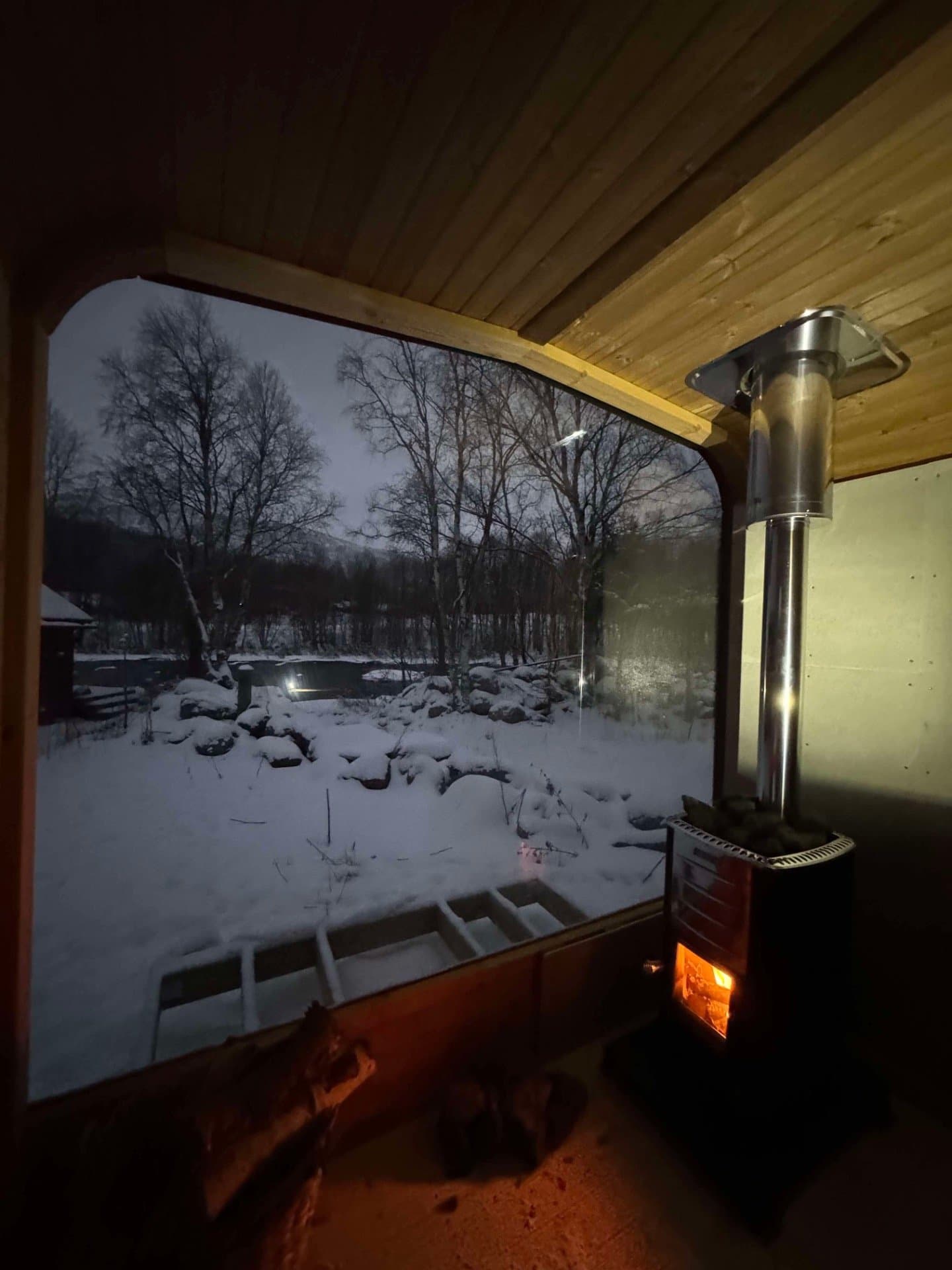 Utsikt fra badstuen gjennom glassfronten mot snødekt hage og bjørketrær i skumringen, med vedovnen tent til høyre