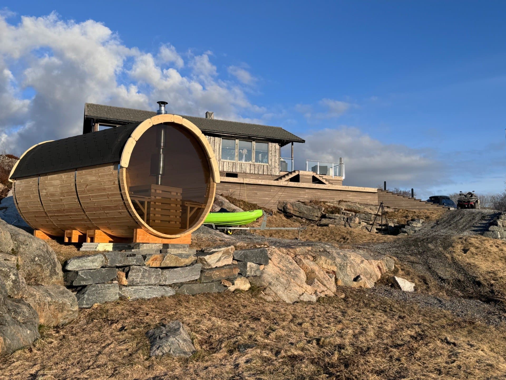 Tønnebadstue med glassvegg i enden på en steinmur i fjæra, med et hus og blå himmel bak