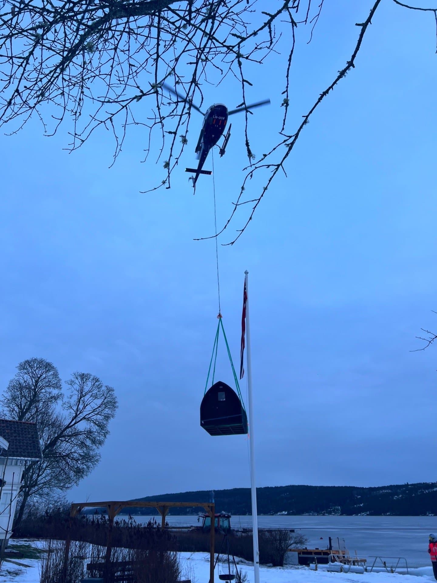 Helikopter som bærer en mørk iglobadstue i tau over en snødekt hage ved et islagt vann