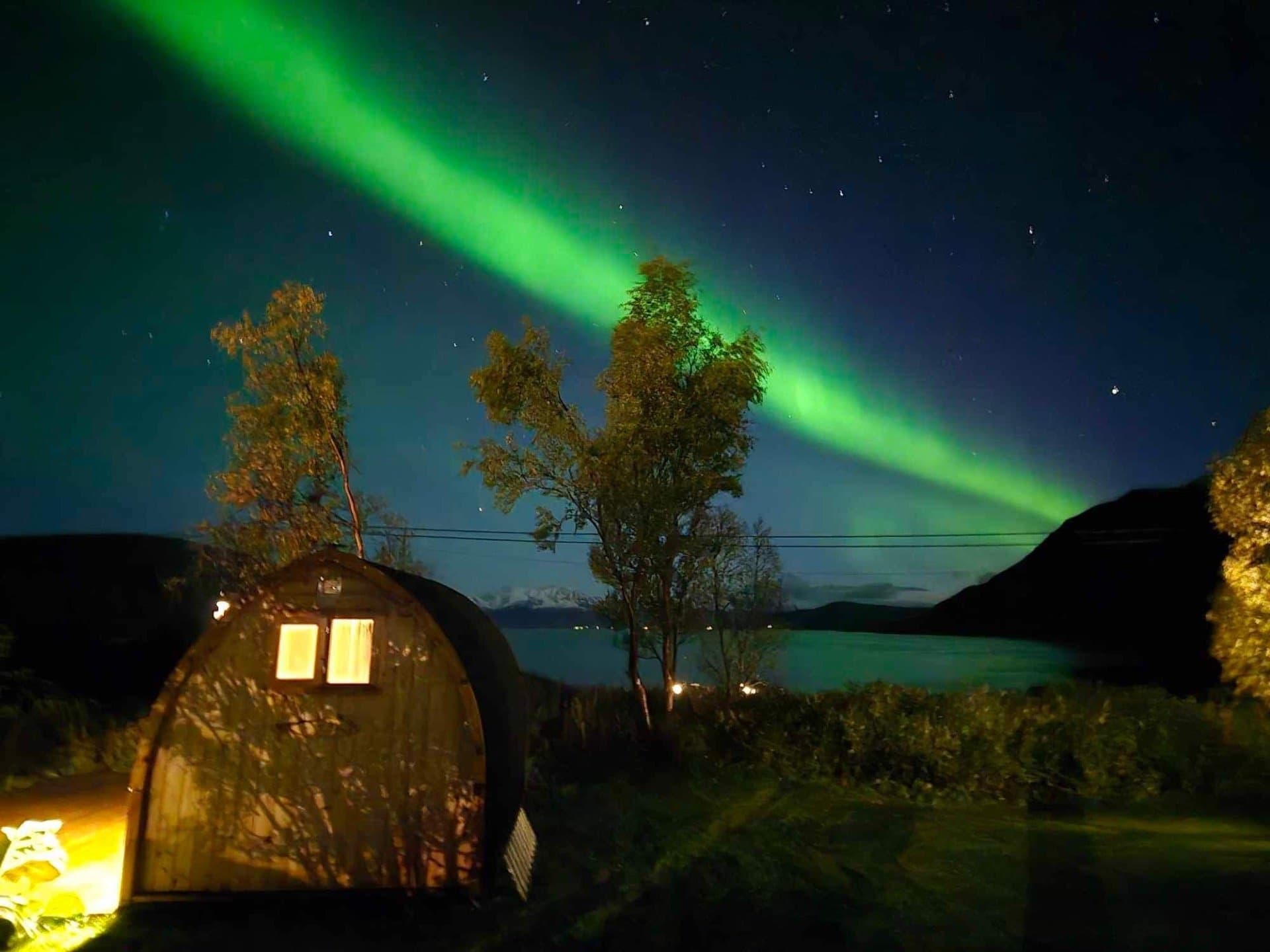 Iglobadstue med opplyste vinduer under grønt nordlys, med fjord og fjell i bakgrunnen