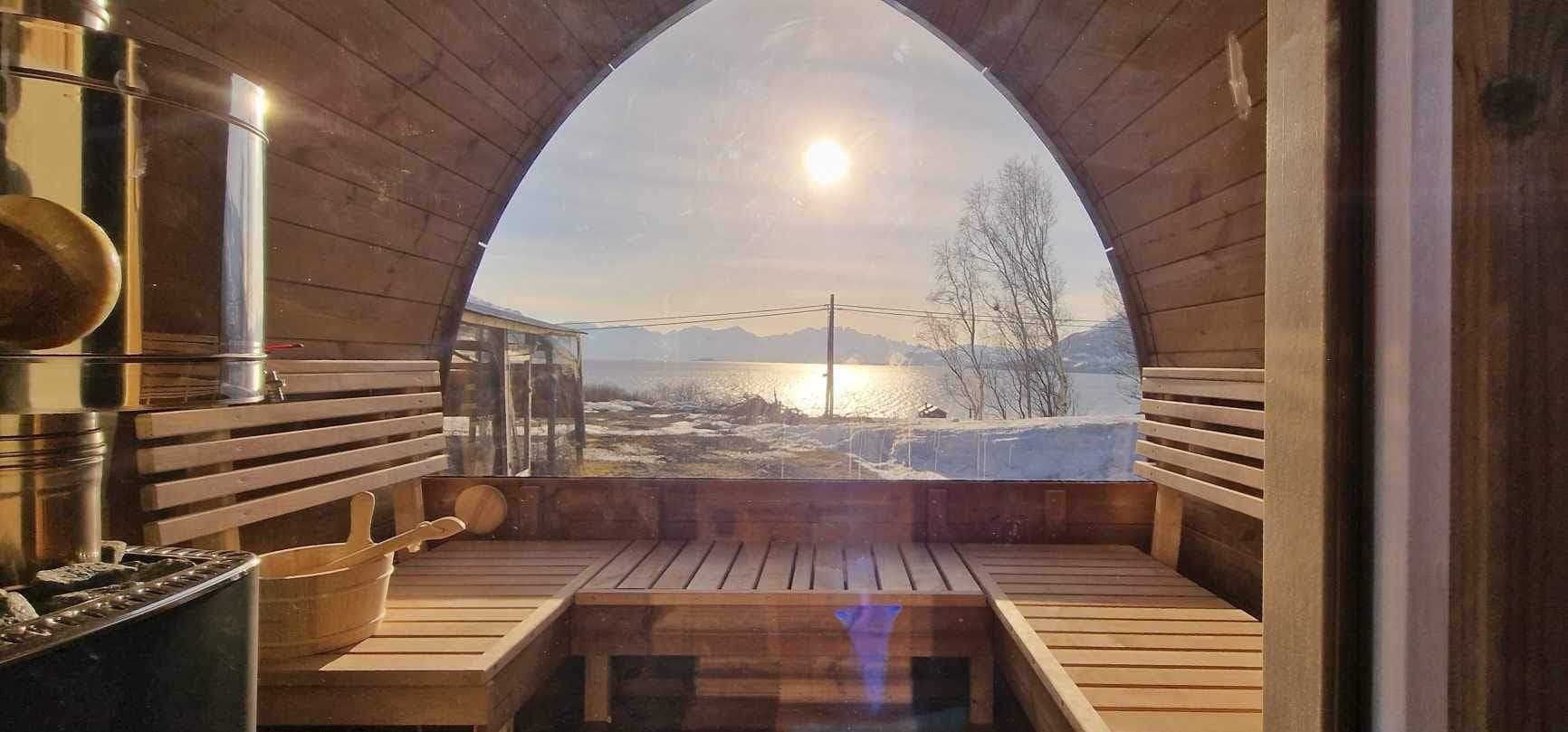 Utsikt fra benkene i en iglobadstue gjennom glassveggen mot sjøen og snødekte fjell i motlys
