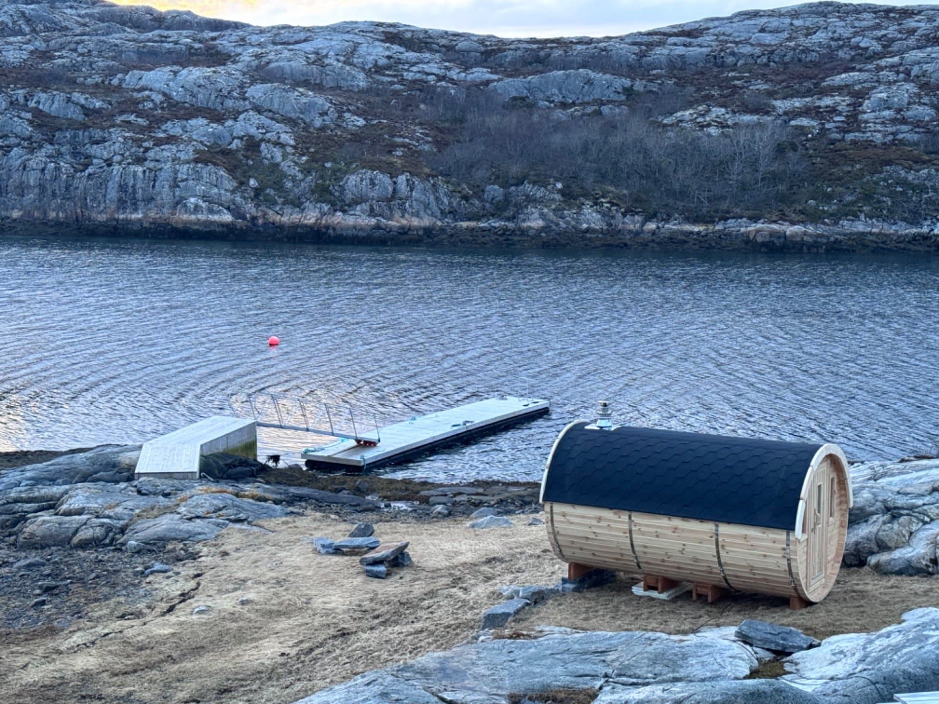 Tønnebadstue på svaberg i fjæra, med en flytebrygge og fjell på andre siden av vannet