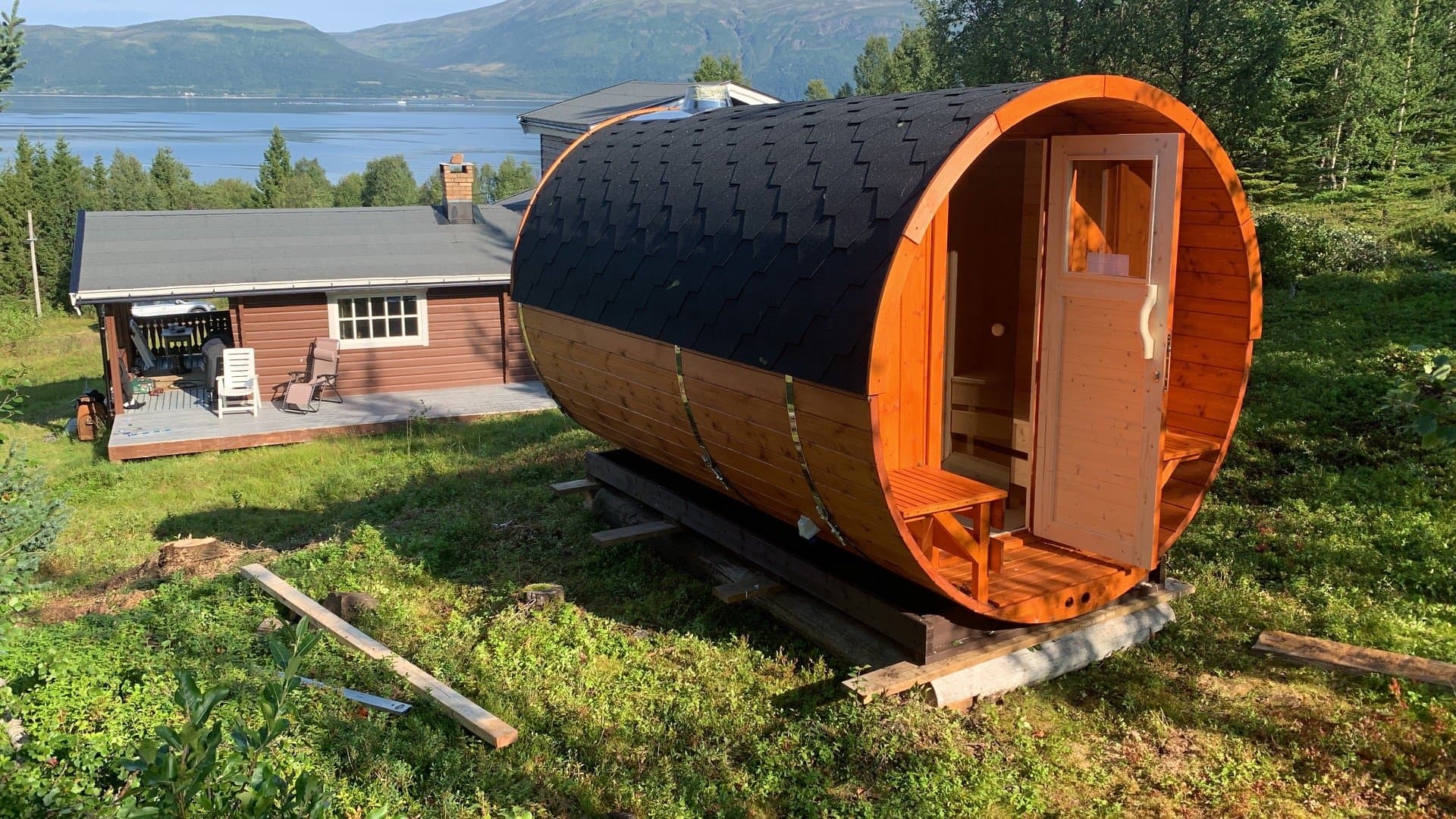 Tønnebadstue på plass på en gressplen ved et hus, med fjorden bak