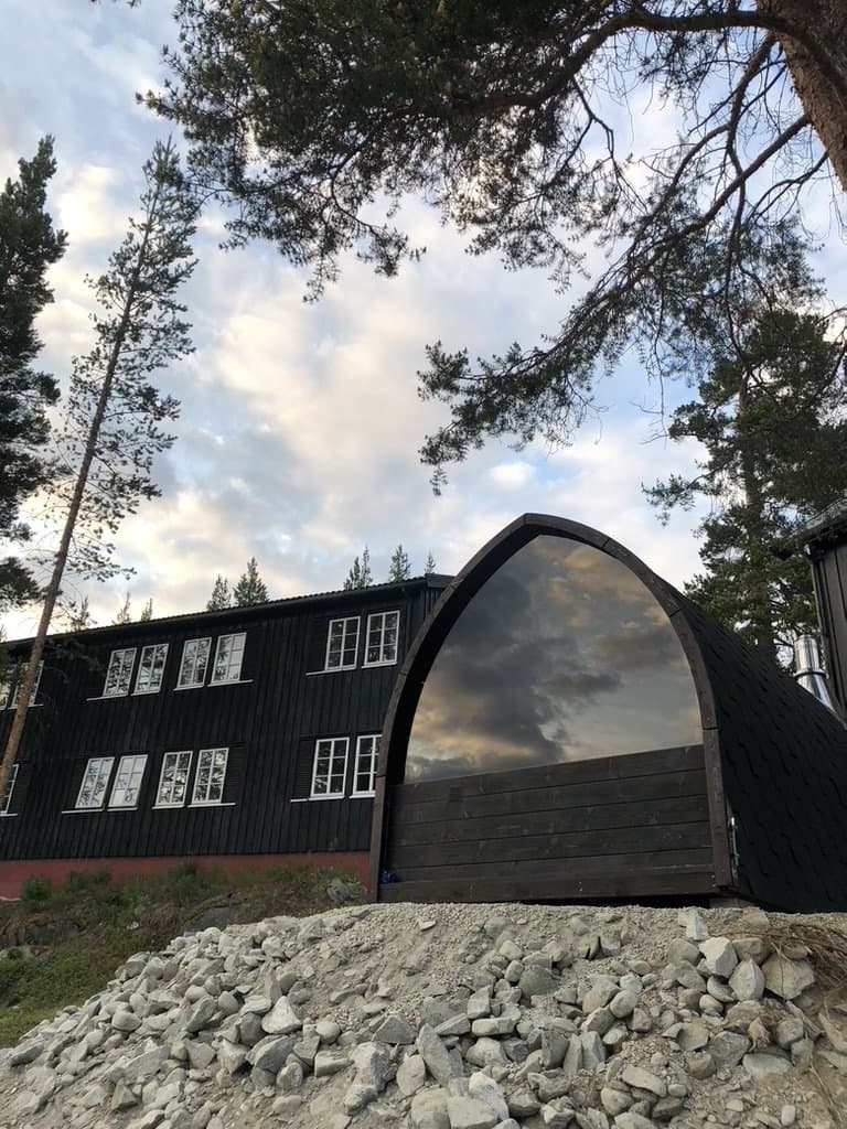 Svart iglobadstue med glassfront ved siden av et mørkt trehus, omgitt av furutrær