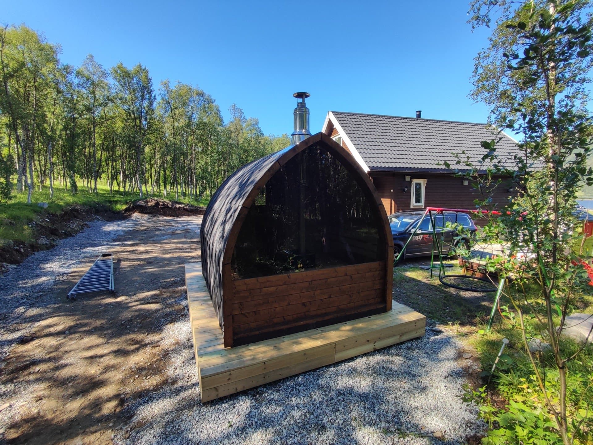 Iglobadstue med pipe på en treplatting på grus, ved et hus og bjørkeskog
