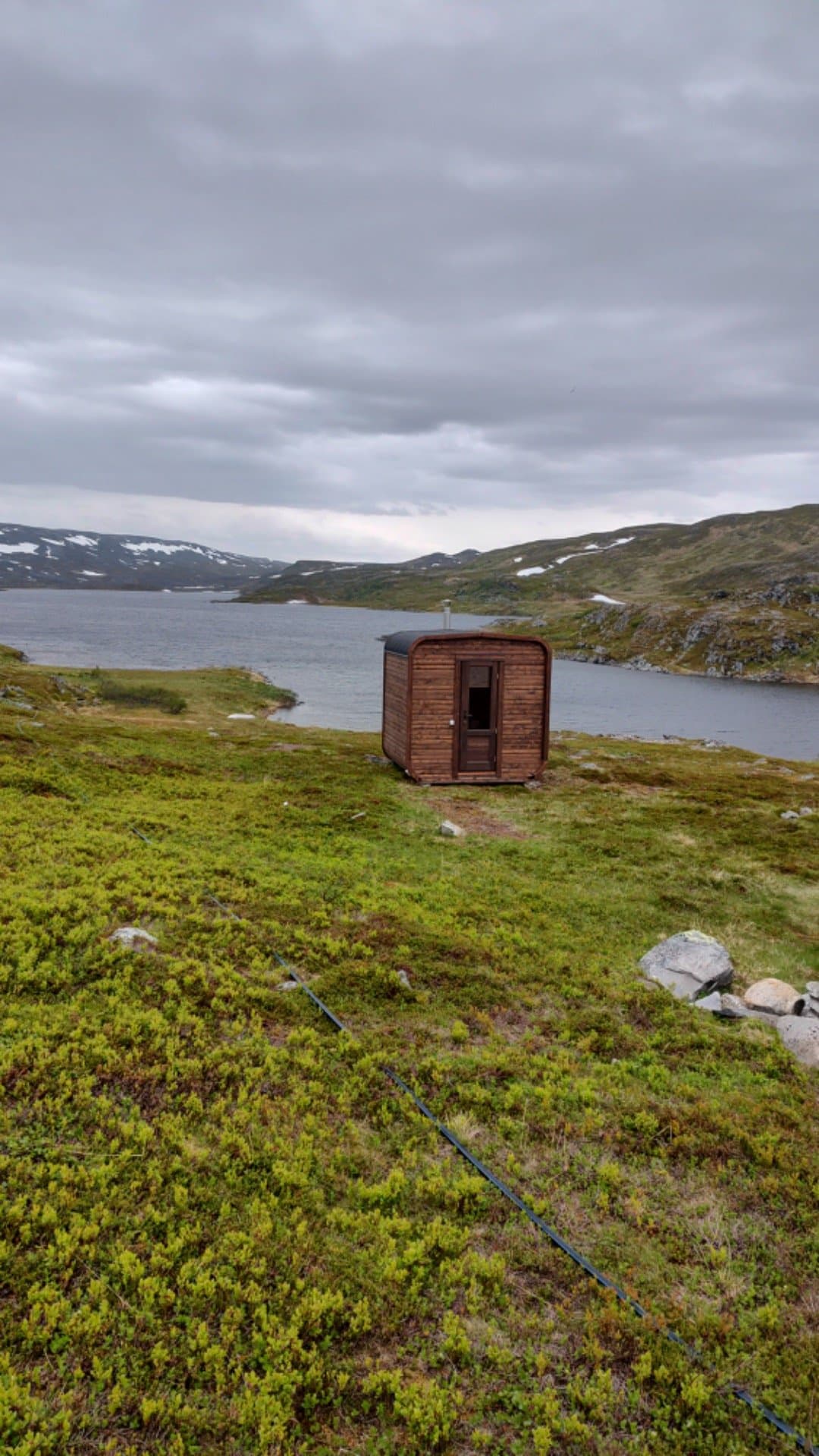 Liten badstue på en lyngslette ved et fjellvann, med snøflekker på fjellene
