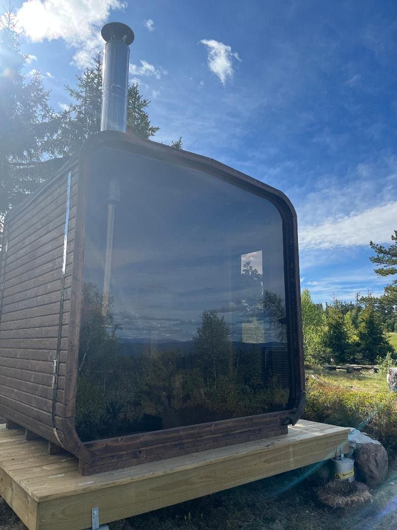 Badstue med stor glassvegg på en platting, med utsikt over vann og skog