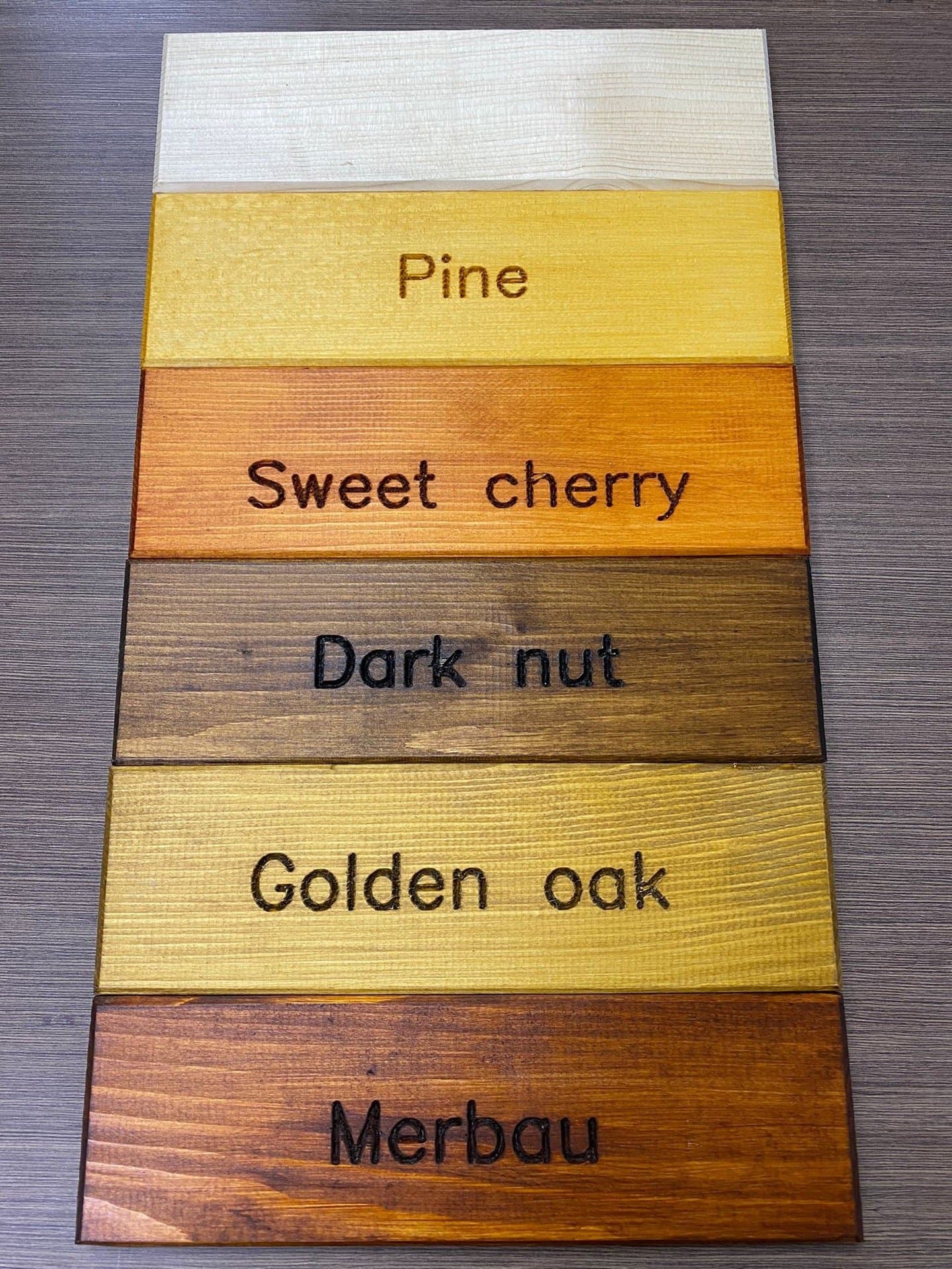 Seks prøvebord med beis i fargene Pine, Sweet cherry, Dark nut, Golden oak og Merbau, og ett ubehandlet